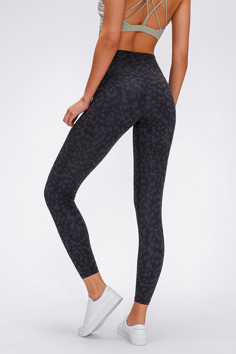 Back view of a model wearing black patterned sports leggings with a wide seamless band waist, fitted full-length silhouette, and one leg bent in a casual pose.