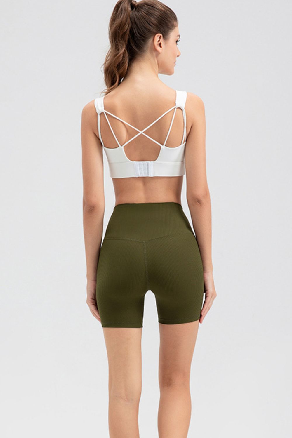 A model poses from the back wearing solid olive-green slim-fit sports shorts with a wide waistband, mid-thigh silhouette, stretch fabric, and center seam.