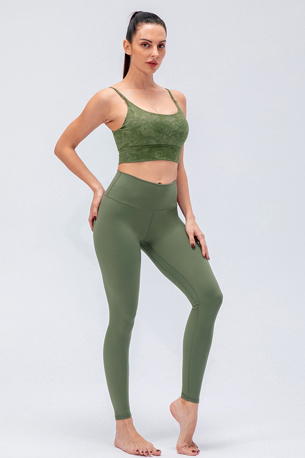 A model poses in solid olive-green active leggings with a wide waistband, slim fit, ankle-length silhouette, and visible front seams.