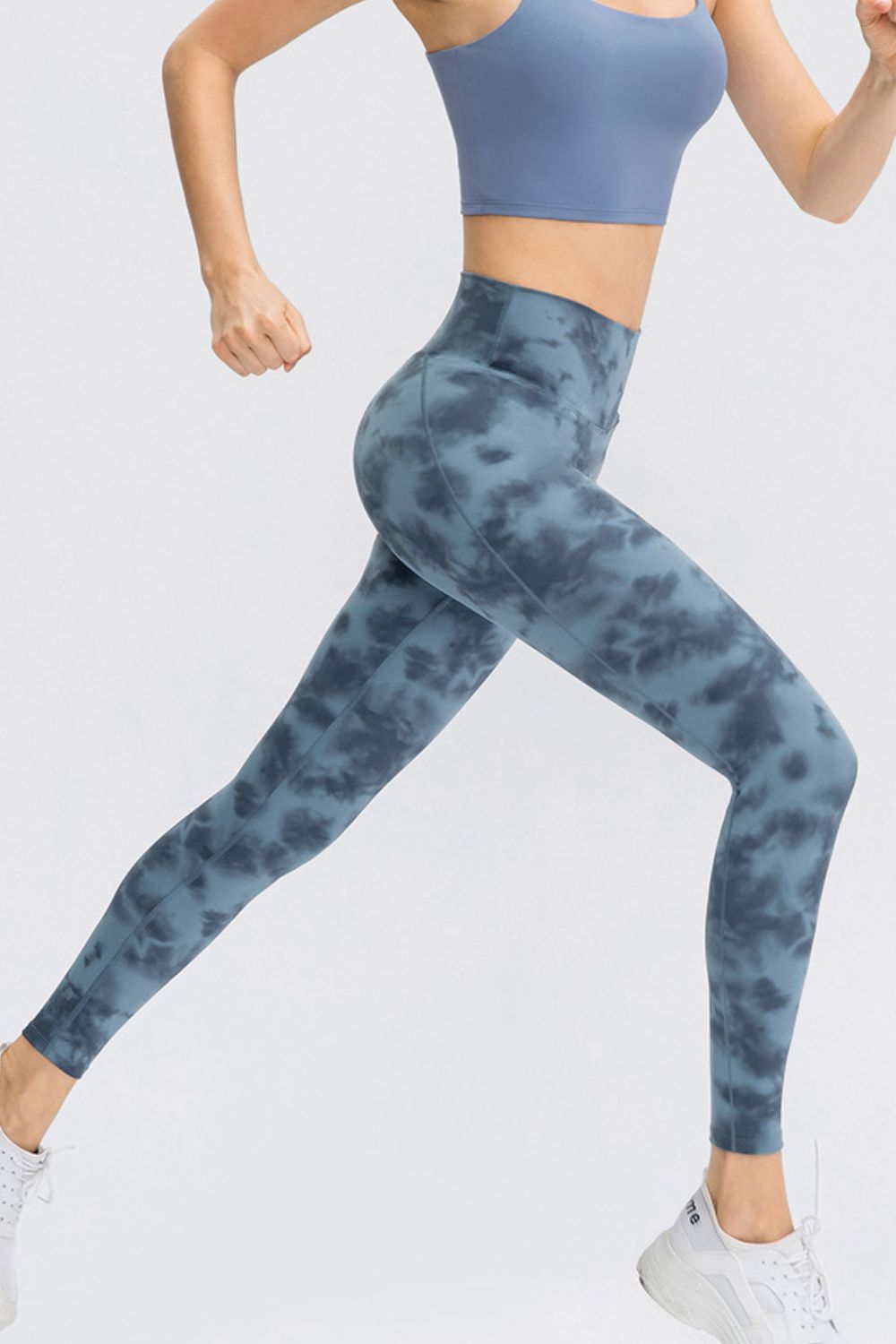 Model in motion wears blue-gray tie-dye leggings with a wide waistband, slim-fit silhouette, long length, and visible curved leg seams.