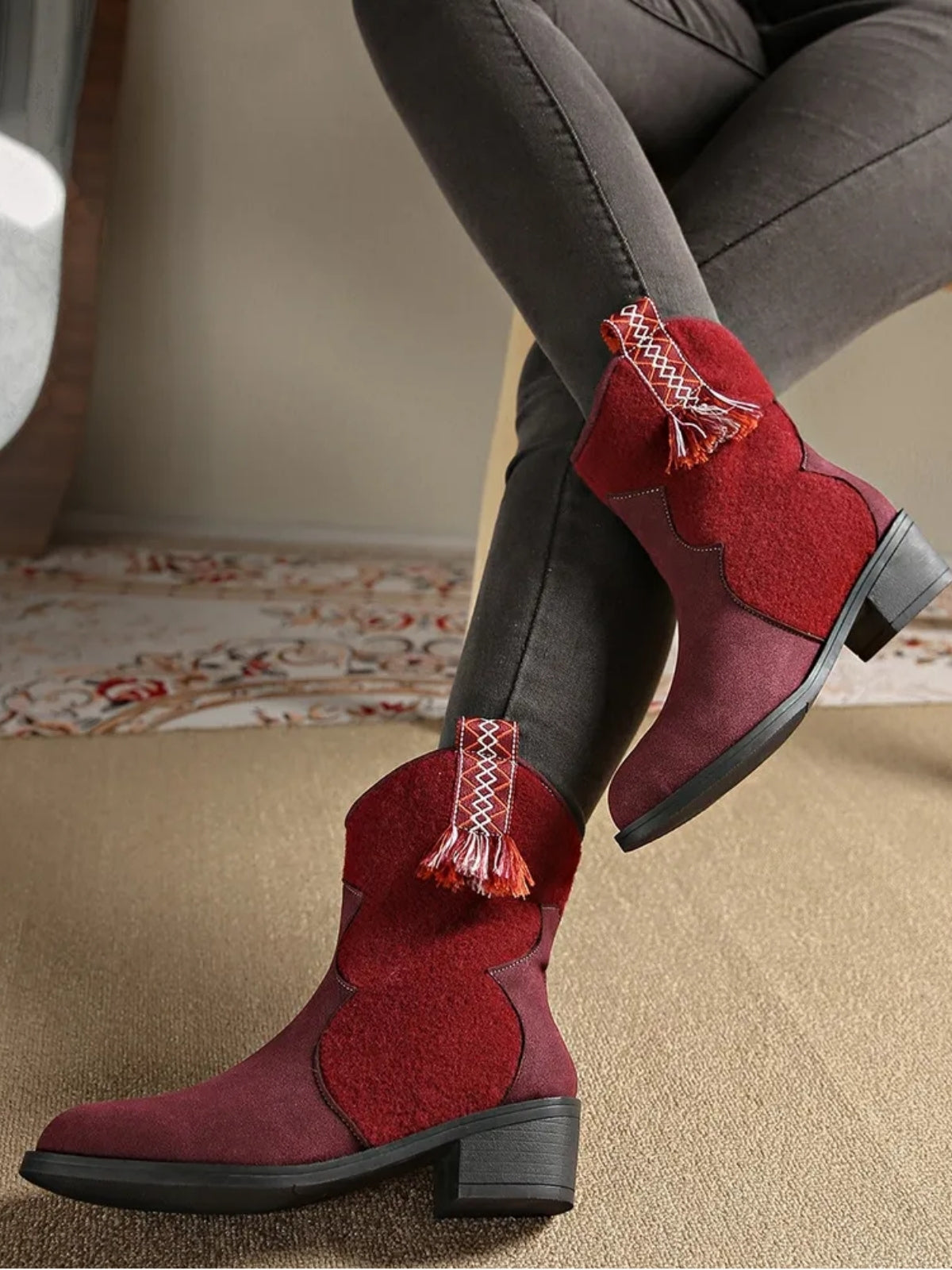 A model crosses her legs wearing burgundy suede point-toe ankle boots with low block heels, elastic side panels, and decorative woven tassel tabs.