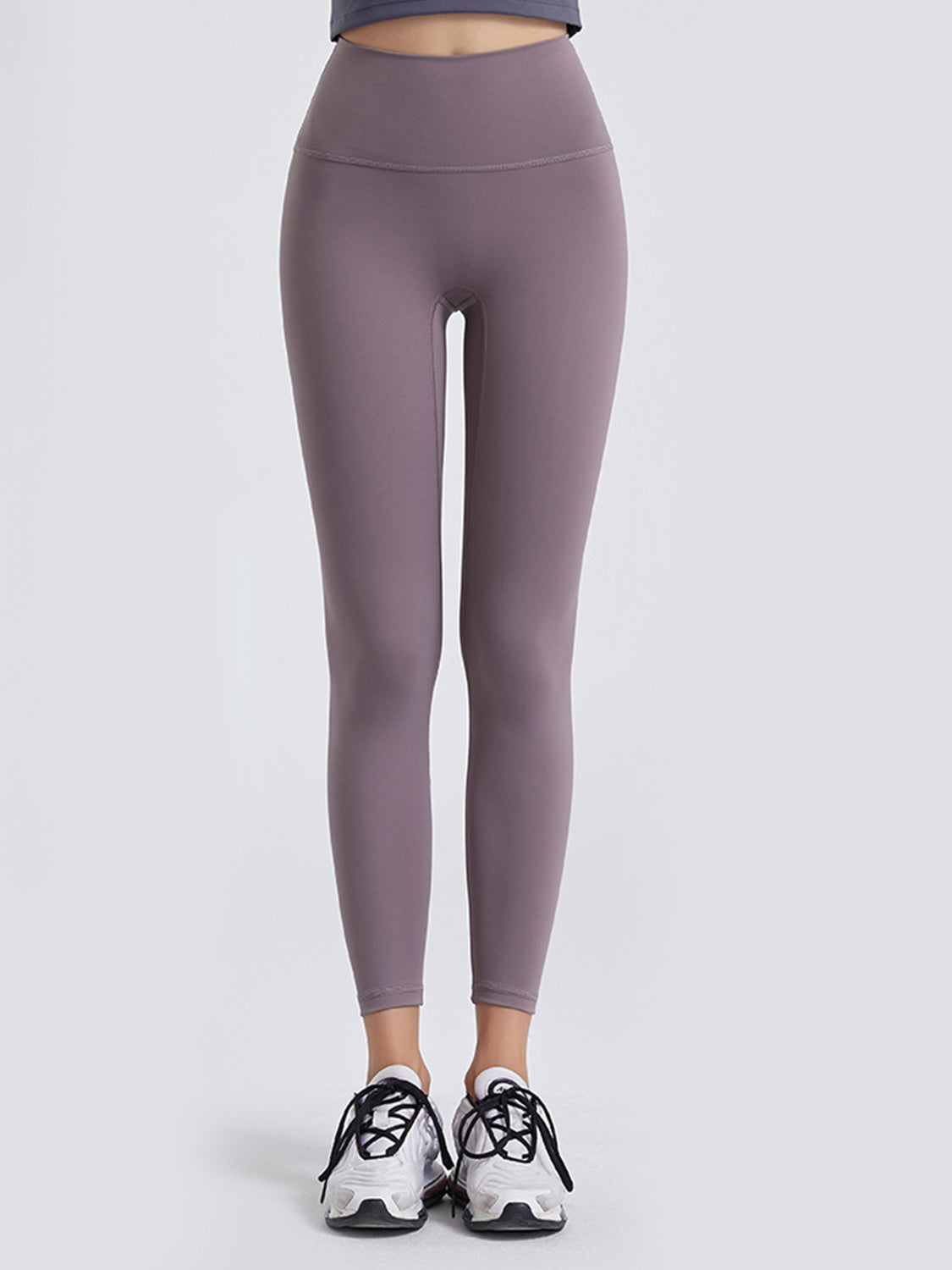 A front-facing model wears mauve wide waistband sports leggings with a high-rise silhouette, smooth stretch construction, and ankle-length tapered legs.