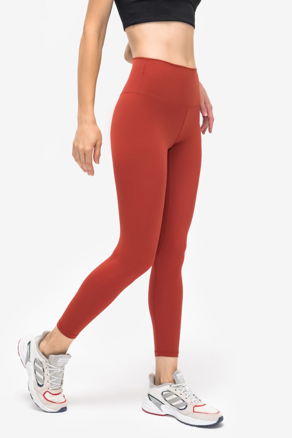 Model poses in rust-red solid sports leggings with a high-rise waistband, slim ankle-length silhouette, and visible seam construction.