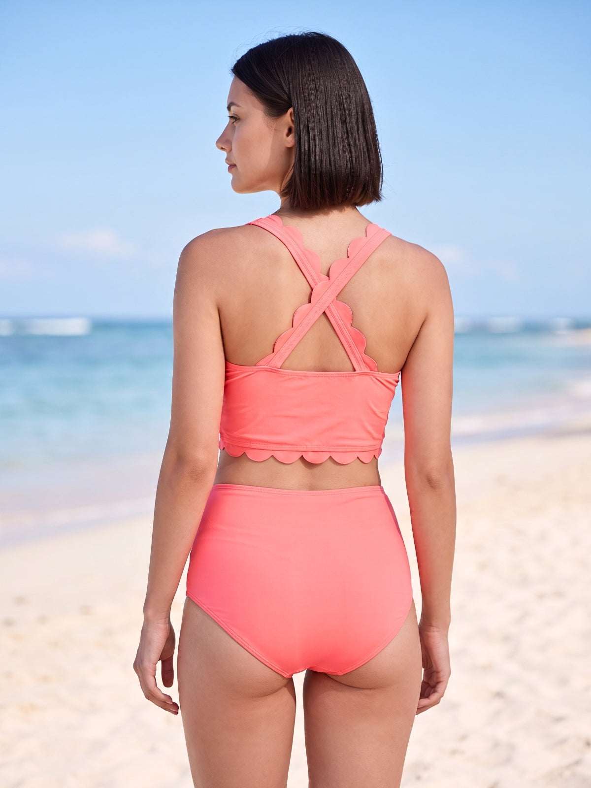 A model faces away wearing a coral two-piece swim set with wide crisscross straps, scalloped trim, a cropped top, and high-waisted bottoms.