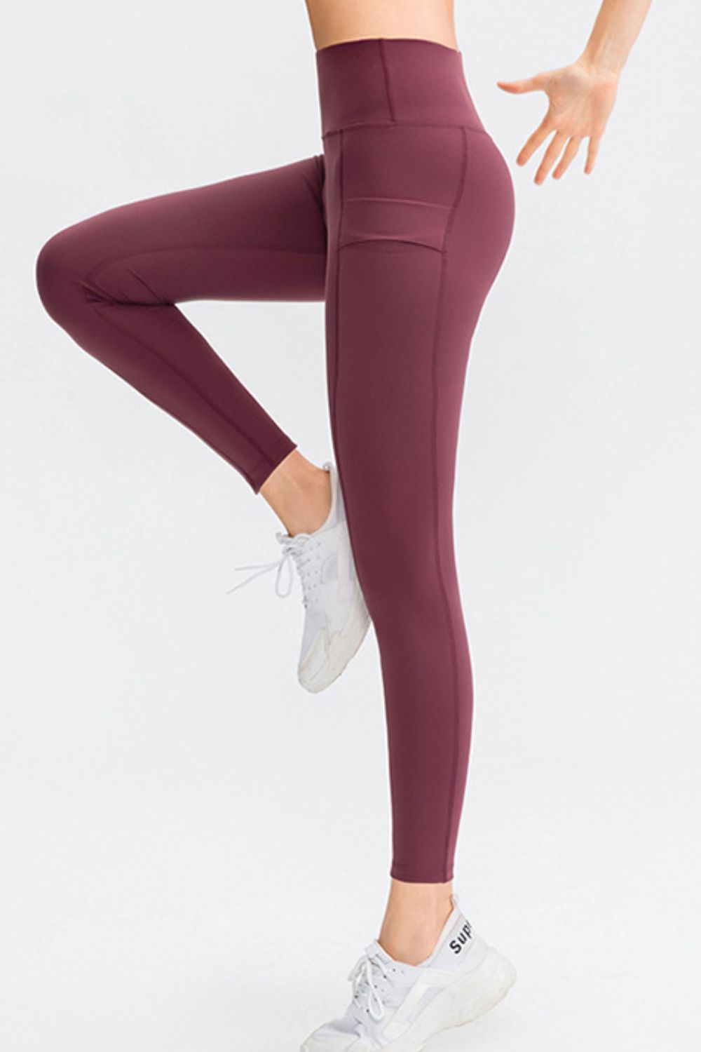 Model balances on one leg in burgundy slim-fit sports leggings with a wide waistband, long tapered silhouette, and visible side pocket.
