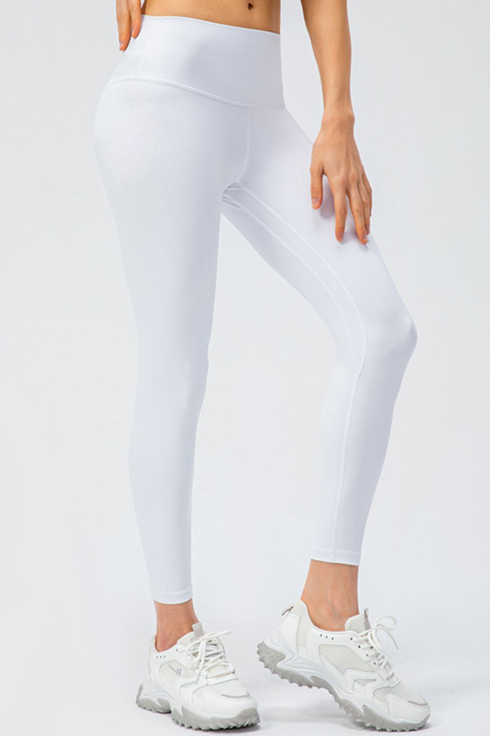 Model wears white wide-waistband slim-fit active leggings with a high-rise silhouette and curved seam detail, standing with one knee bent.