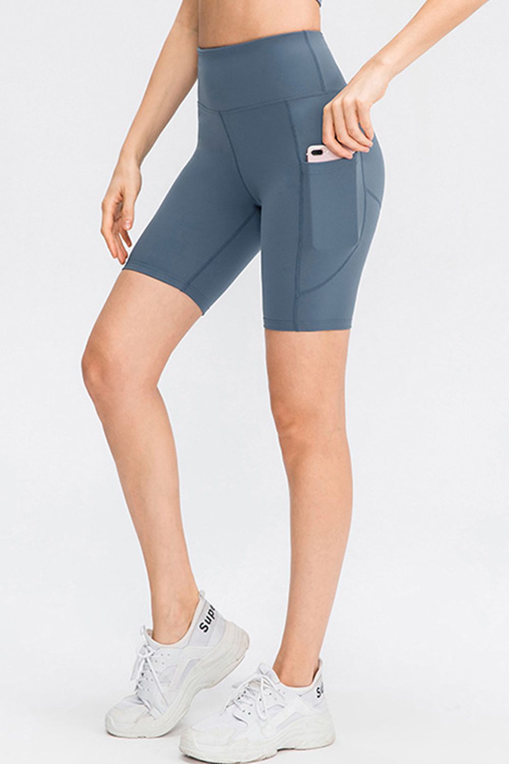 Model poses in slate blue wide-waistband sports shorts with a fitted mid-thigh silhouette, visible seam construction, and a side pocket holding a phone.