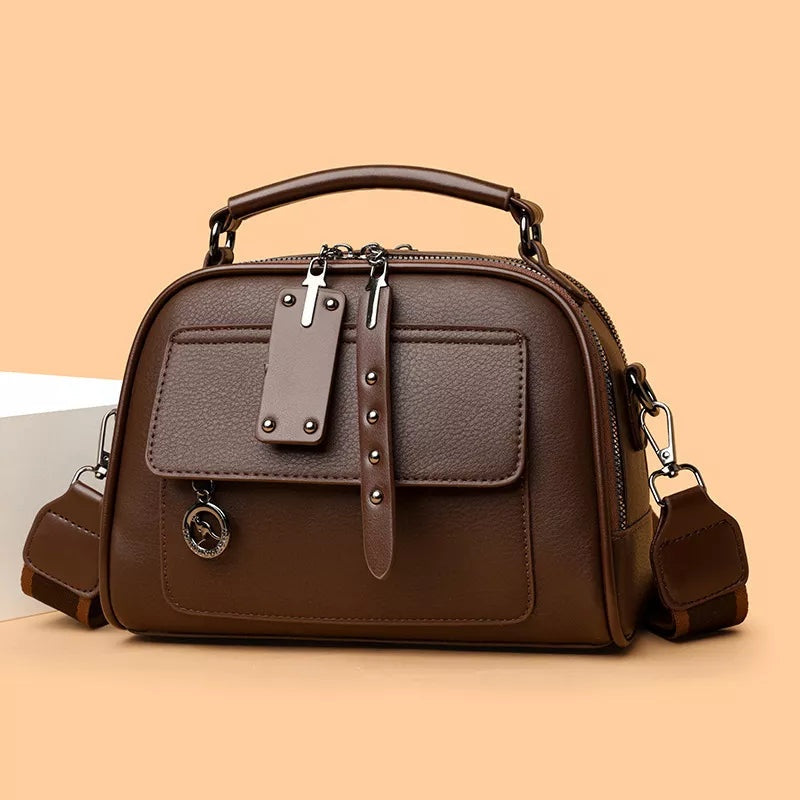 Small brown PU leather crossbody bag with an adjustable strap, top handle, dual zip closure, front pockets, and structured compact silhouette.