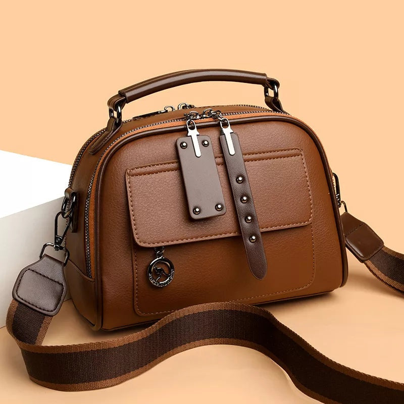 Small brown PU leather crossbody bag with an adjustable woven strap, top handle, structured silhouette, front pocket, and dual zipper closures.