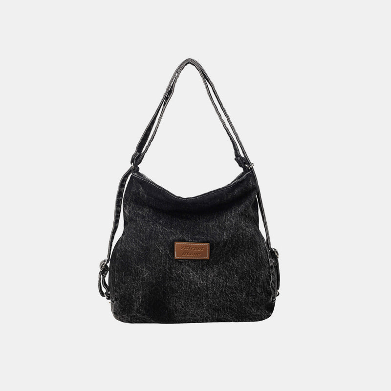 Black denim shoulder bag with a softly slouching silhouette, gently shaped base, adjustable strap, and centered logo patch.