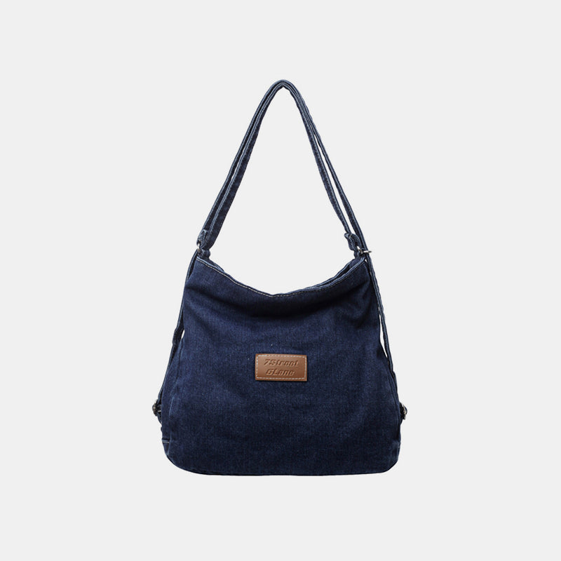 Dark blue denim shoulder bag with an adjustable strap, softly slouching silhouette, gently shaped base, and a subtle front logo patch.