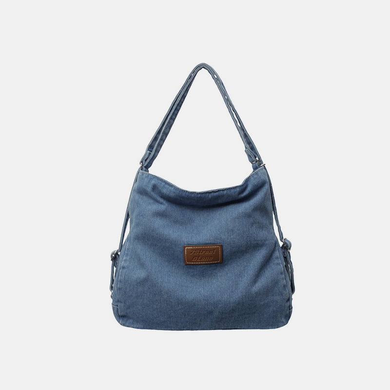 Blue denim shoulder bag with an adjustable strap, softly slouching silhouette, gently shaped base, and a subtle centered logo patch.