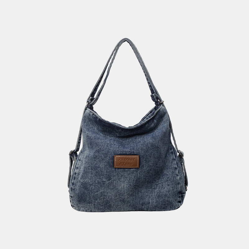 A medium blue denim shoulder bag features an adjustable strap, softly slouching silhouette, gently shaped base, and centered logo patch.
