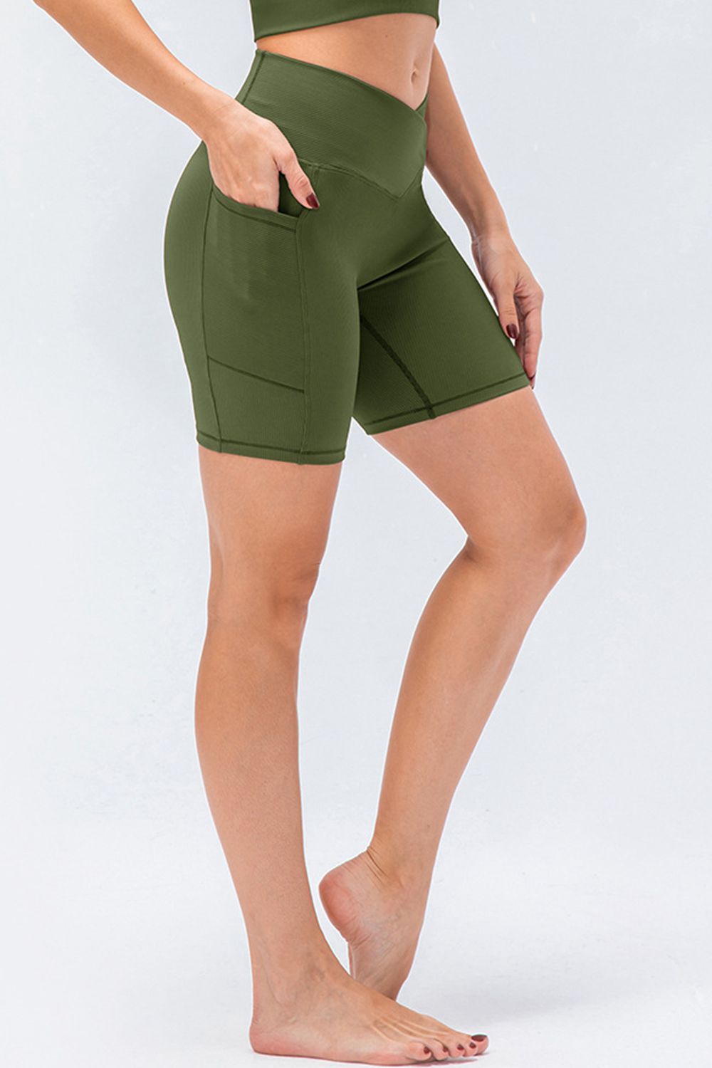 A model wears olive green slim-fit V-waistband sports shorts with above-knee length, side pocket, and panel seams, posing with one hand tucked in.