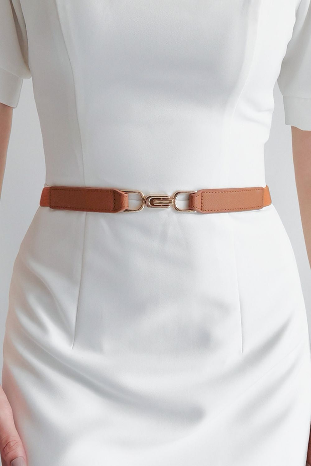 Model wears a slim brown elastic belt over a white dress, featuring stitched edges and a sculptural interlocking alloy buckle that defines the waist.