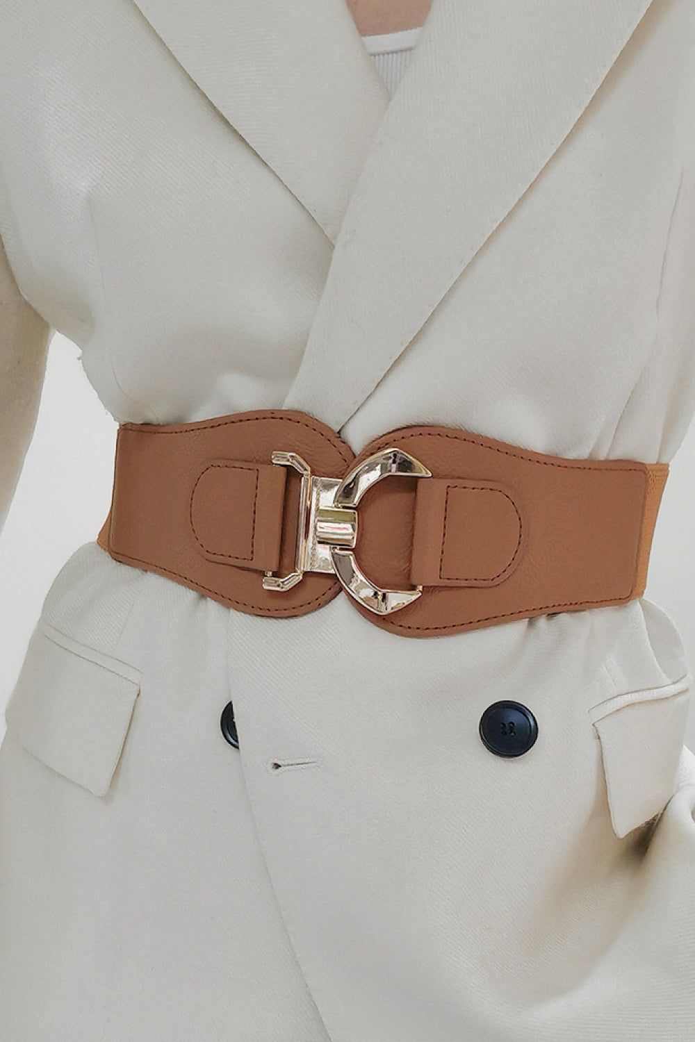 A model wears a broad brown elastic belt with curved ends, stitched PU construction, and a sculptural silver alloy buckle over a white tailored outfit.