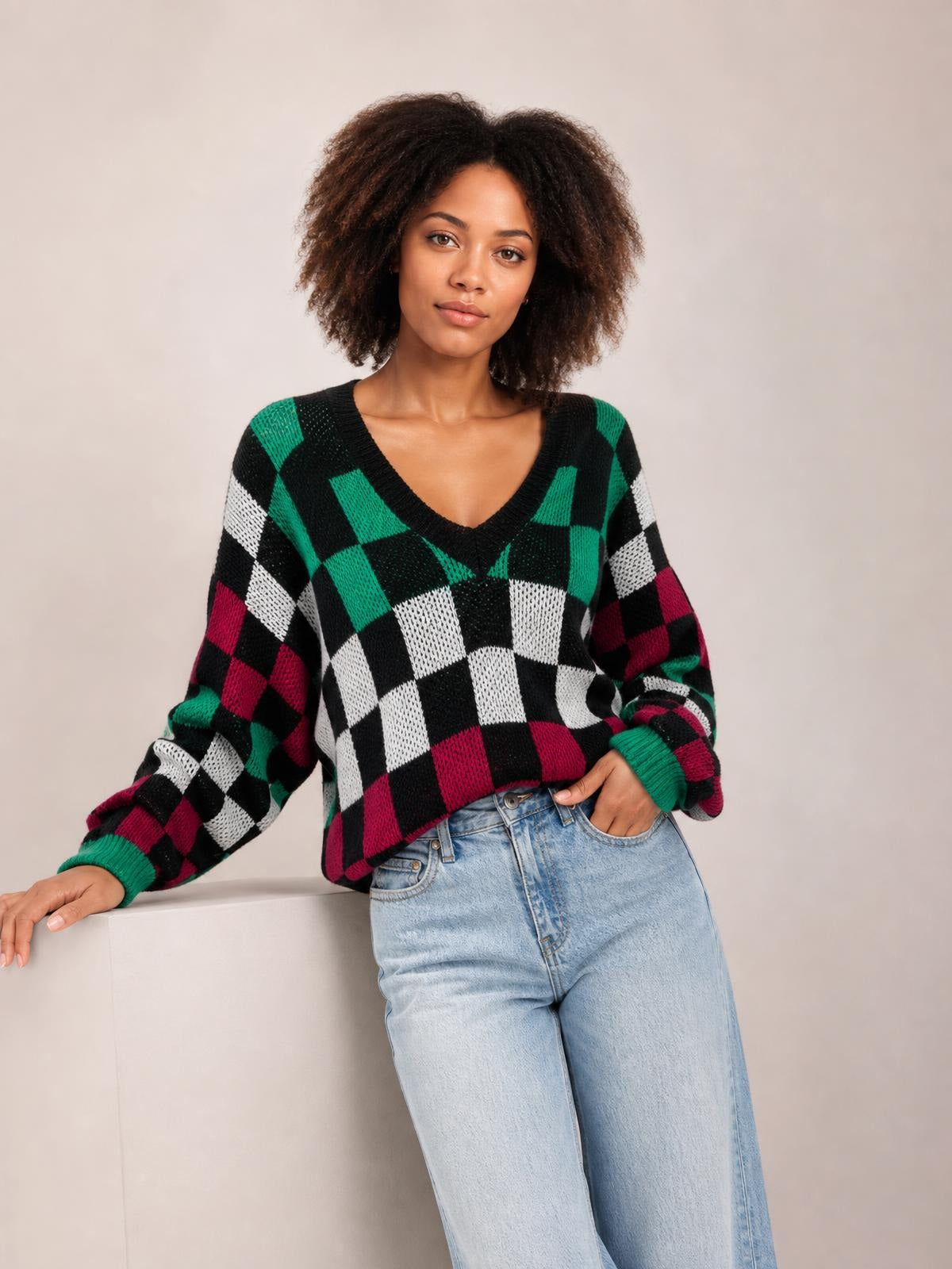 Model wears a relaxed checkered V-neck sweater in black, green, white, and burgundy with voluminous lantern sleeves and ribbed cuffs, leaning with one hand in her jeans pocket.