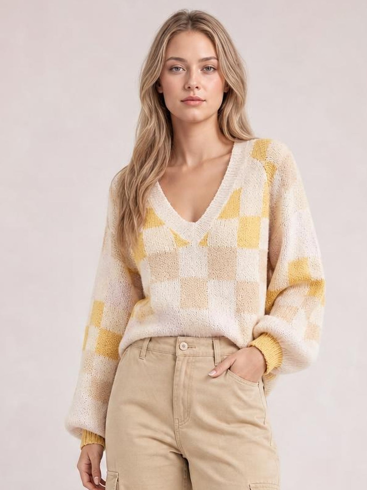 Model wears a cream and mustard checkered V-neck sweater with relaxed lantern sleeves and ribbed cuffs, posing with one hand in her trouser pocket.