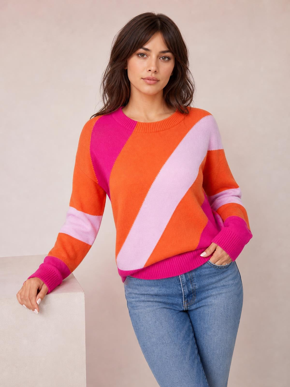Model wears a bright orange, pink, and lavender color block ribbed sweater with a round neck, long sleeves, and regular silhouette while posing with one hand in her jeans pocket.