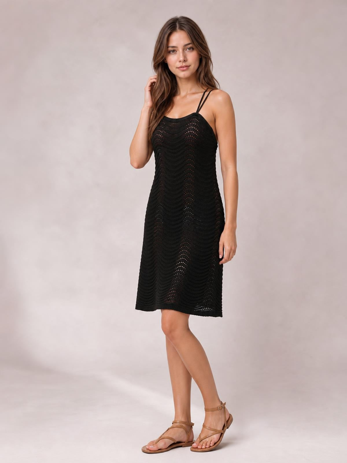 A model poses with one hand in her hair, wearing a black sleeveless openwork cover-up dress with a straight neckline, slim silhouette, and knee-length hem.