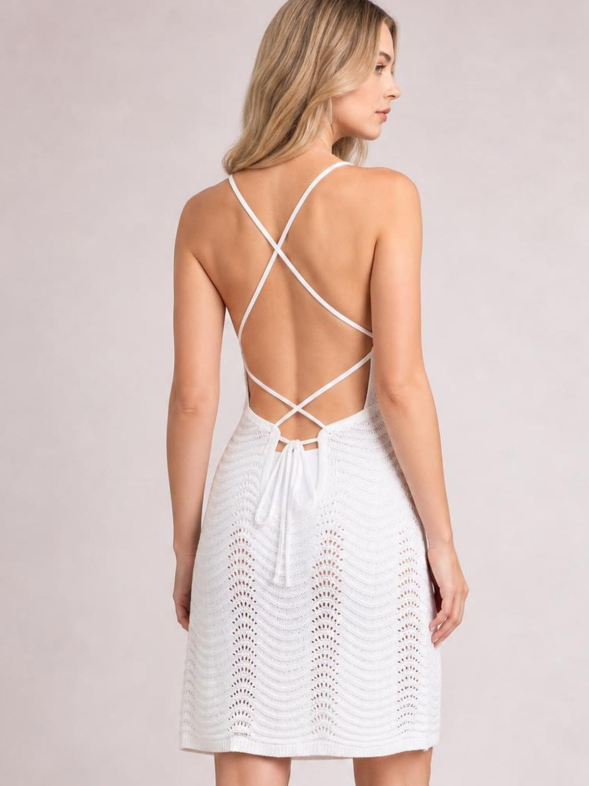 Model viewed from behind wearing a white crisscross halter neck cover-up dress with a backless design, sheer openwork knit, tie waist, and relaxed mini silhouette.