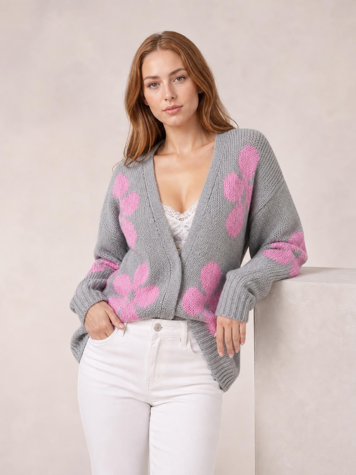 Model wears a gray open-front long-sleeve cardigan with pink flower motifs, chunky knit texture, ribbed cuffs, and a relaxed silhouette, posing with one hand in her pocket.
