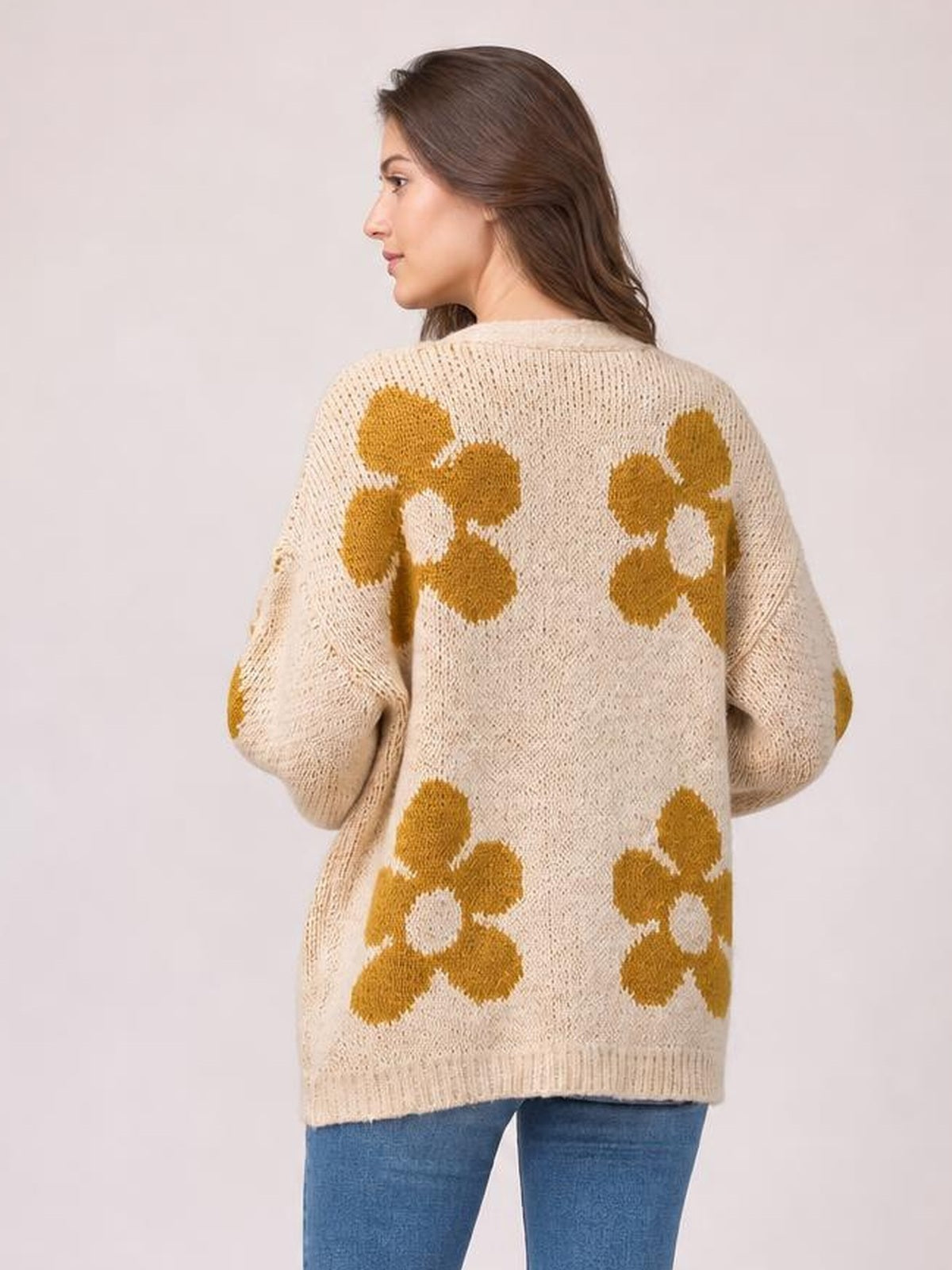 A model turns to show the cream knit cardigan’s relaxed long-sleeve silhouette and bold mustard flower motifs across the back.