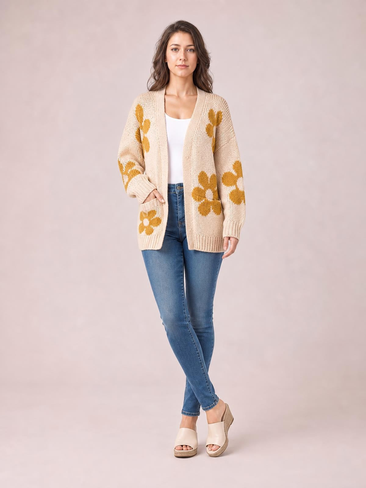 A woman poses in a beige open-front long-sleeve cardigan with mustard flower motifs, ribbed cuffs and hem, front pockets, and a relaxed silhouette.