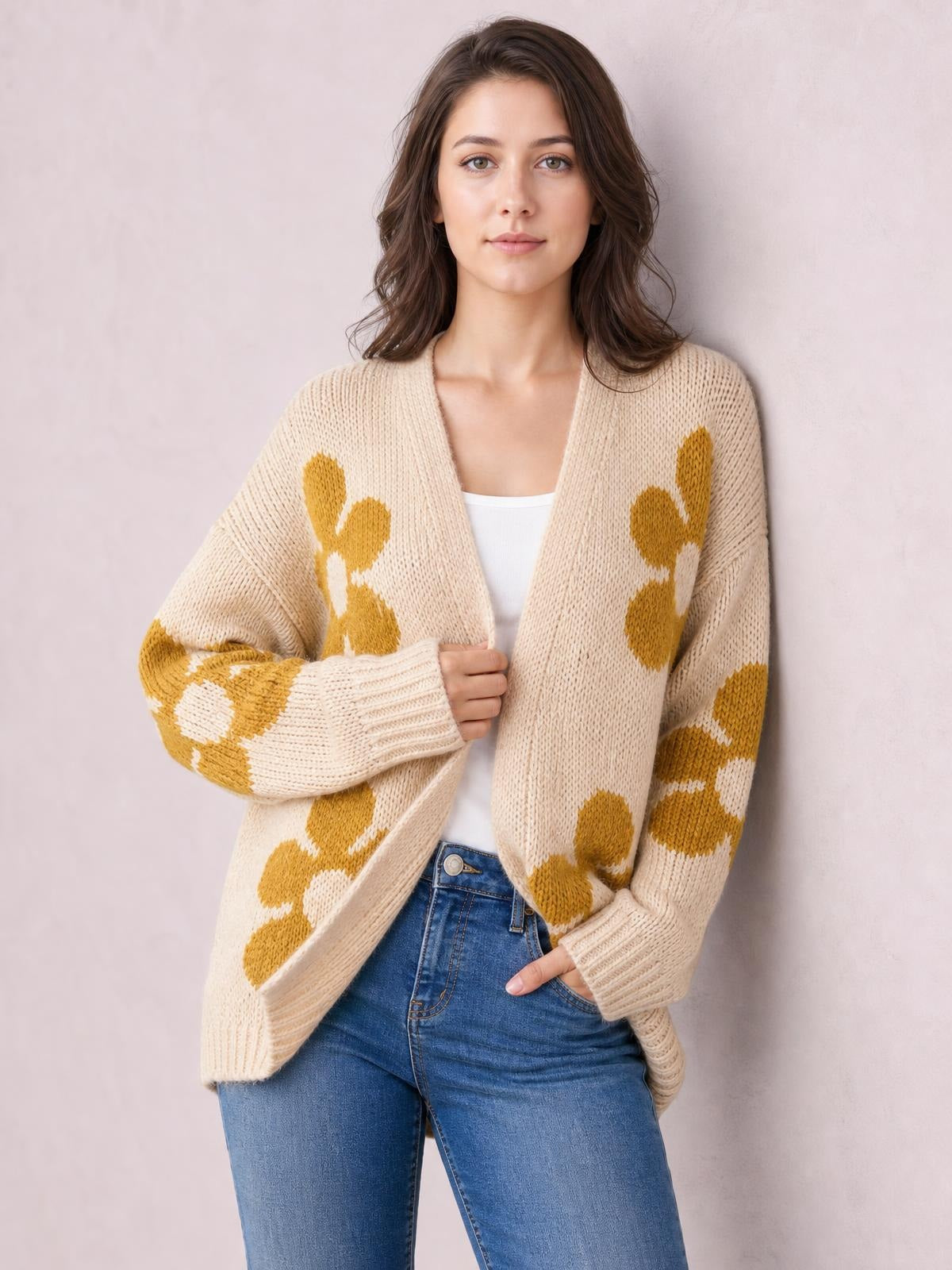 Model wearing a beige open-front long-sleeve cardigan with mustard flower motifs, relaxed knit silhouette, ribbed cuffs and hem, over a white top and jeans.