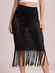 Model wears a black openwork cover-up skirt with a drawstring waist, straight midi silhouette, and long fringe hem with tassel details.