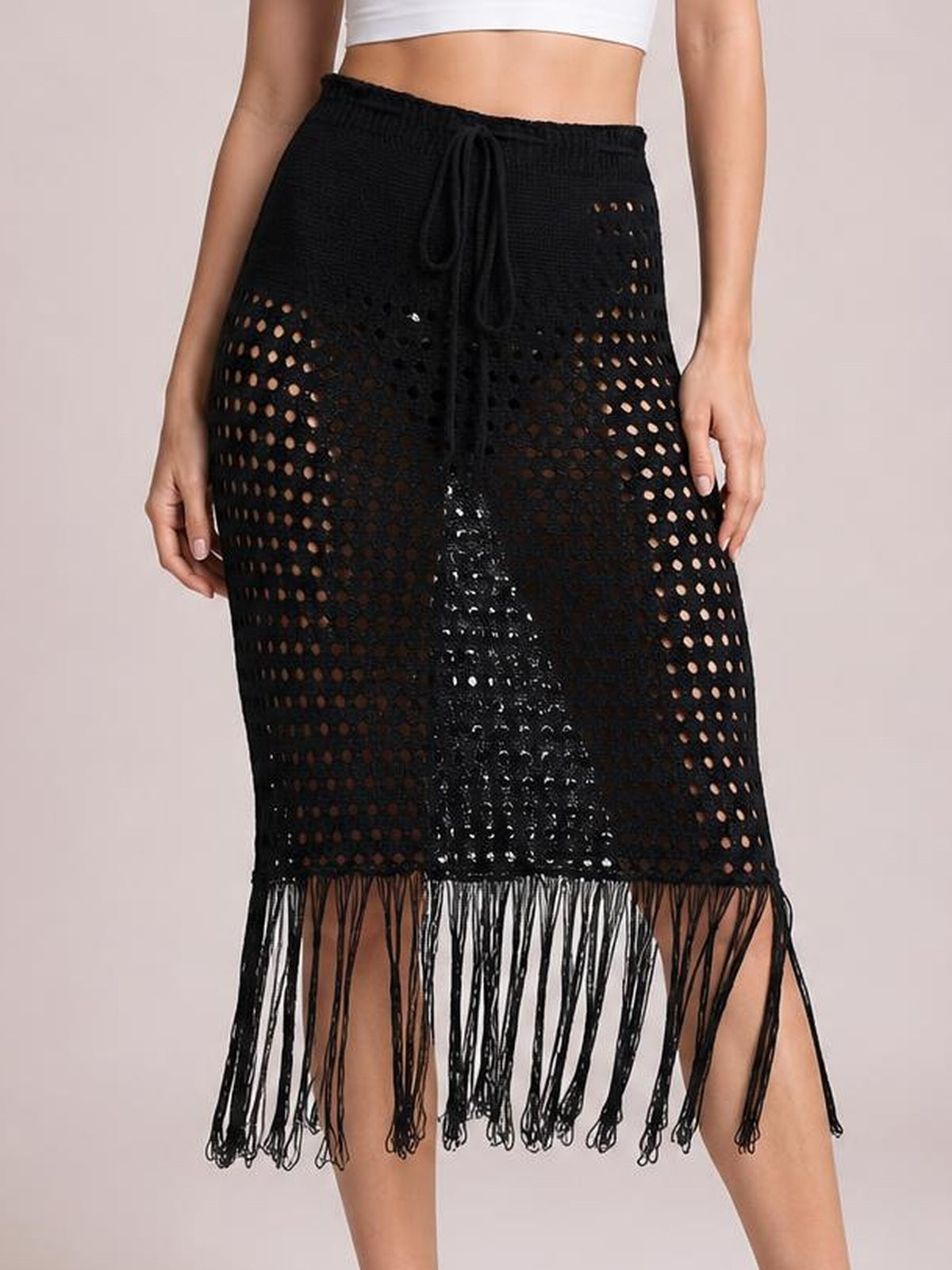 Model wears a black openwork cover-up skirt with a drawstring waist, straight midi silhouette, and long fringe hem with tassel details.