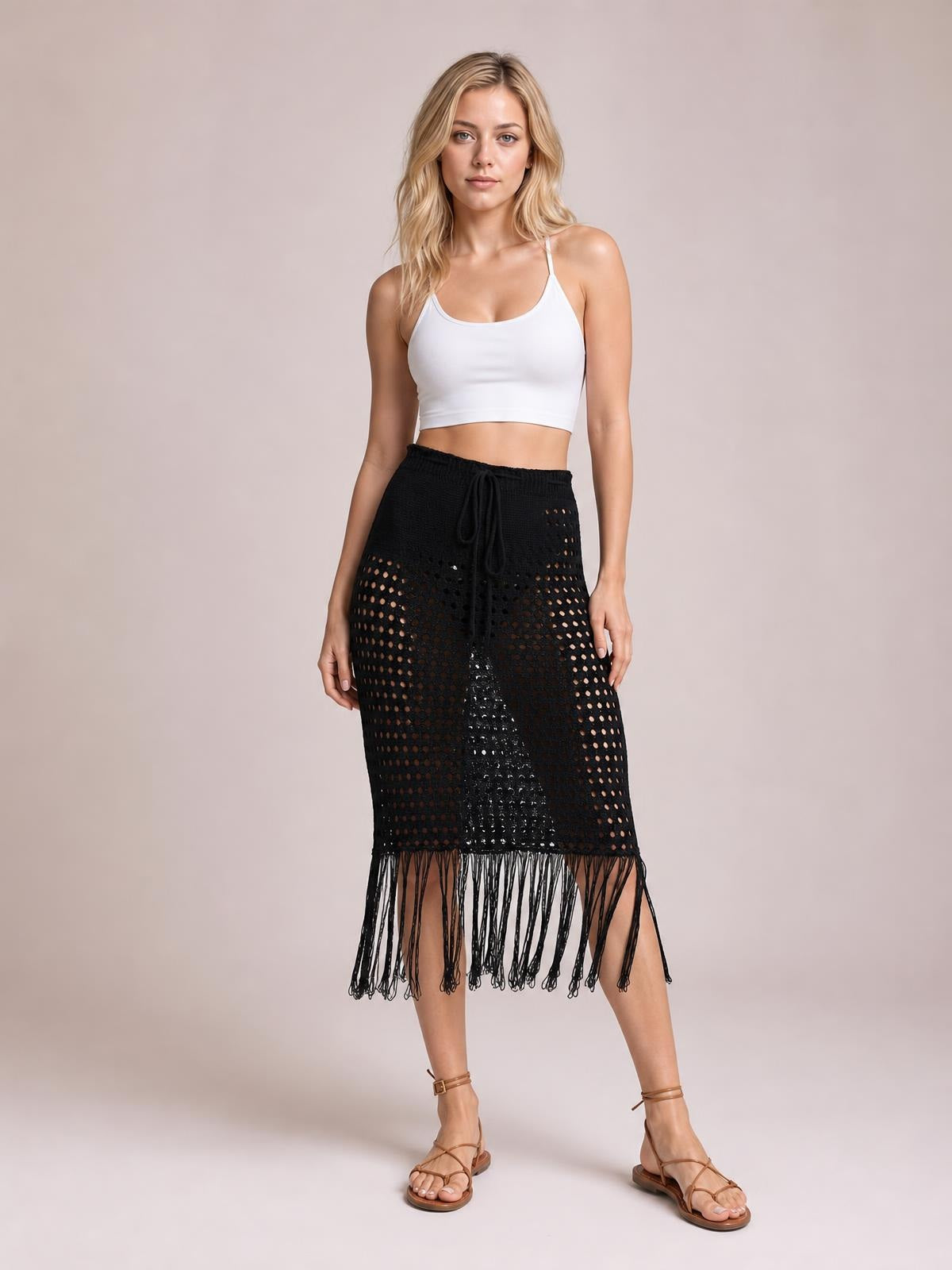 Model stands in a black openwork cover-up skirt with a drawstring waist, straight silhouette, and long fringe hem, styled with a white crop top.