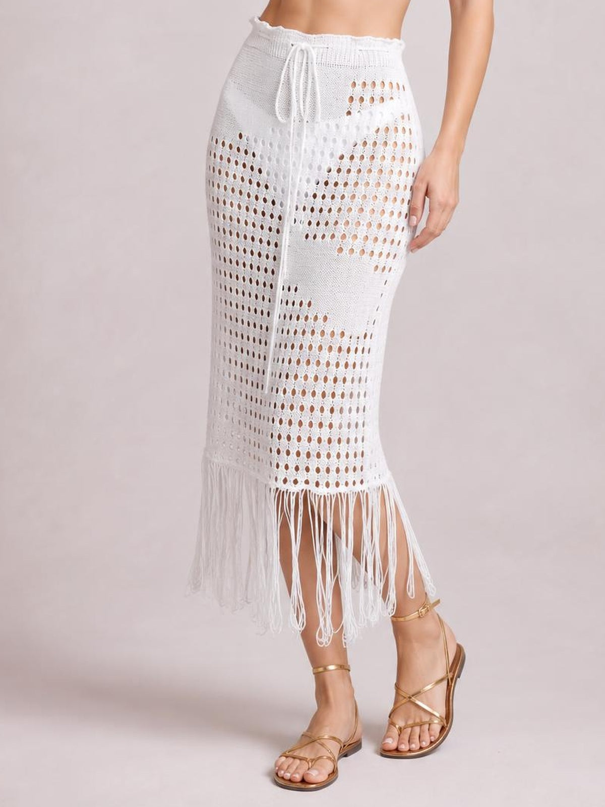 Model wears a white openwork midi skirt with a drawstring waist, straight silhouette, patterned knit construction, and long fringe hem while standing with one leg crossed.