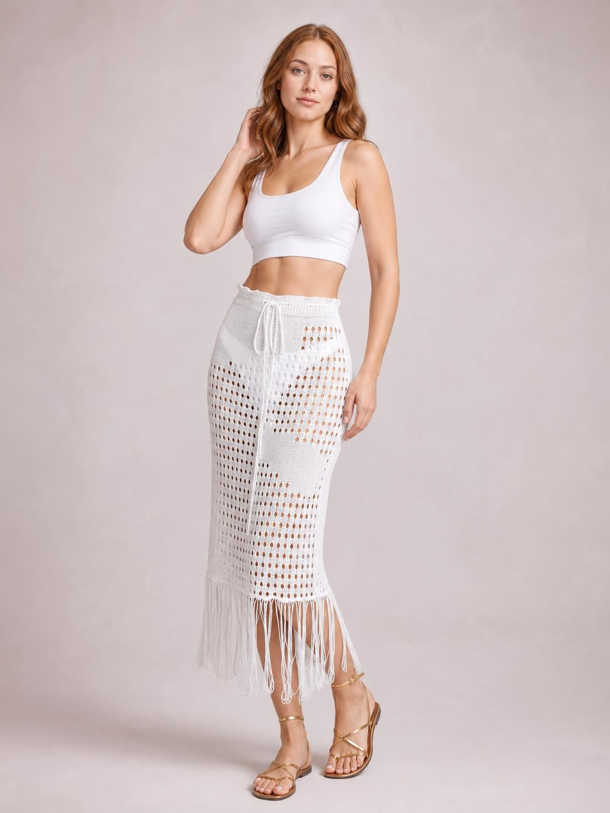 Model poses in a white openwork cover-up skirt with a drawstring waist, fitted midi silhouette, and long fringe hem.