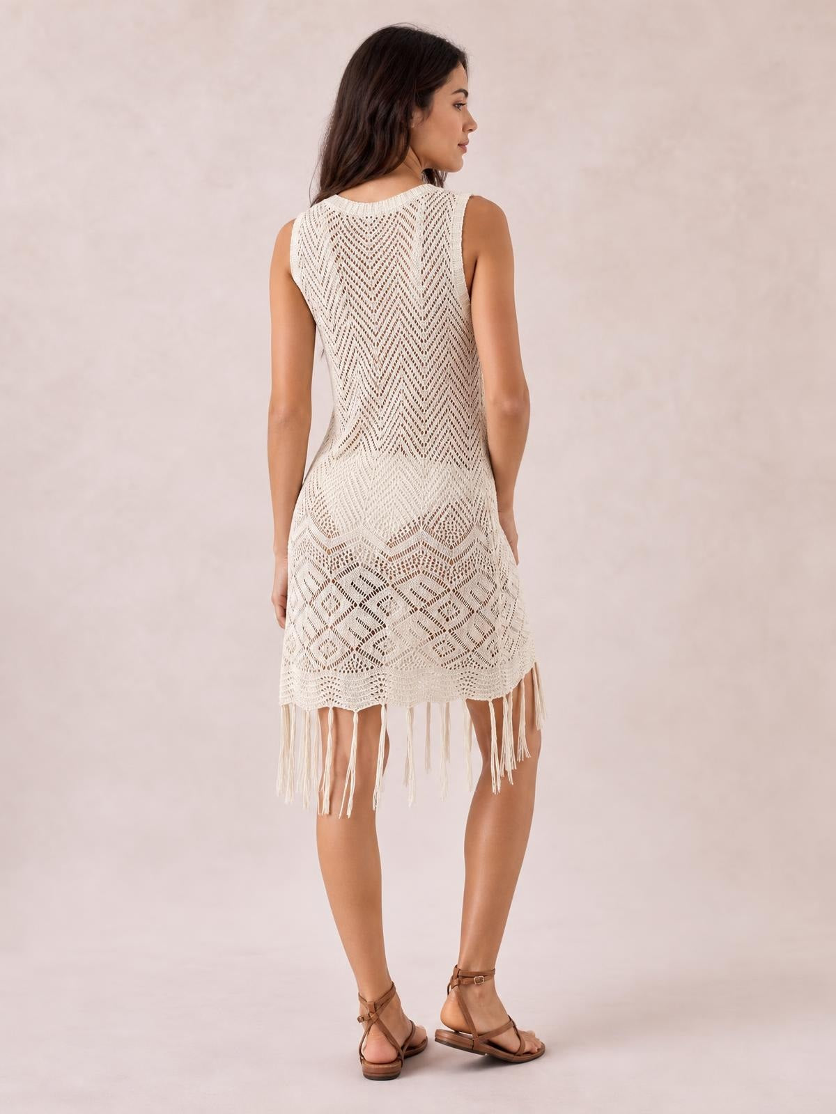 Model shown from the back in a cream sleeveless openwork cover-up dress with a fitted silhouette, geometric knit pattern, and long fringe trim hem.