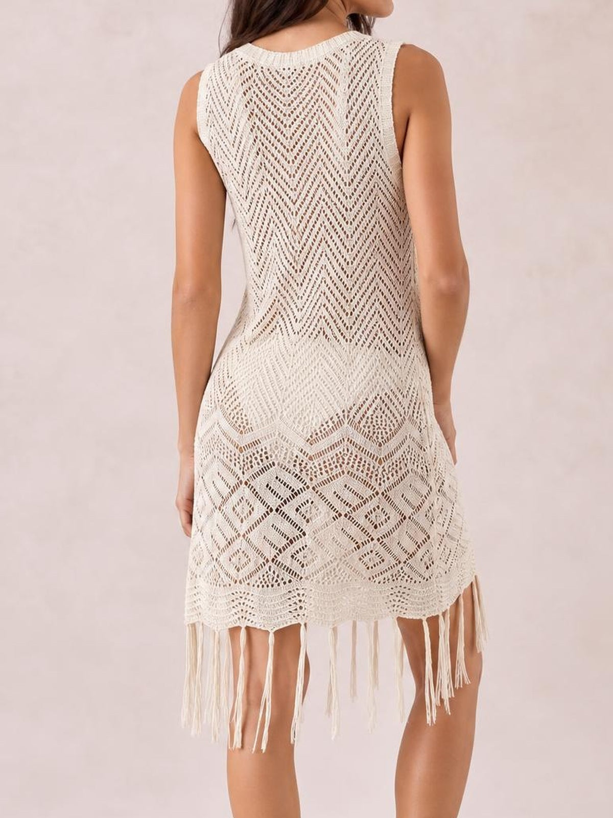 A model stands turned away in a cream sleeveless openwork cover-up dress with a relaxed silhouette, geometric knit pattern, and long tassel fringe hem.