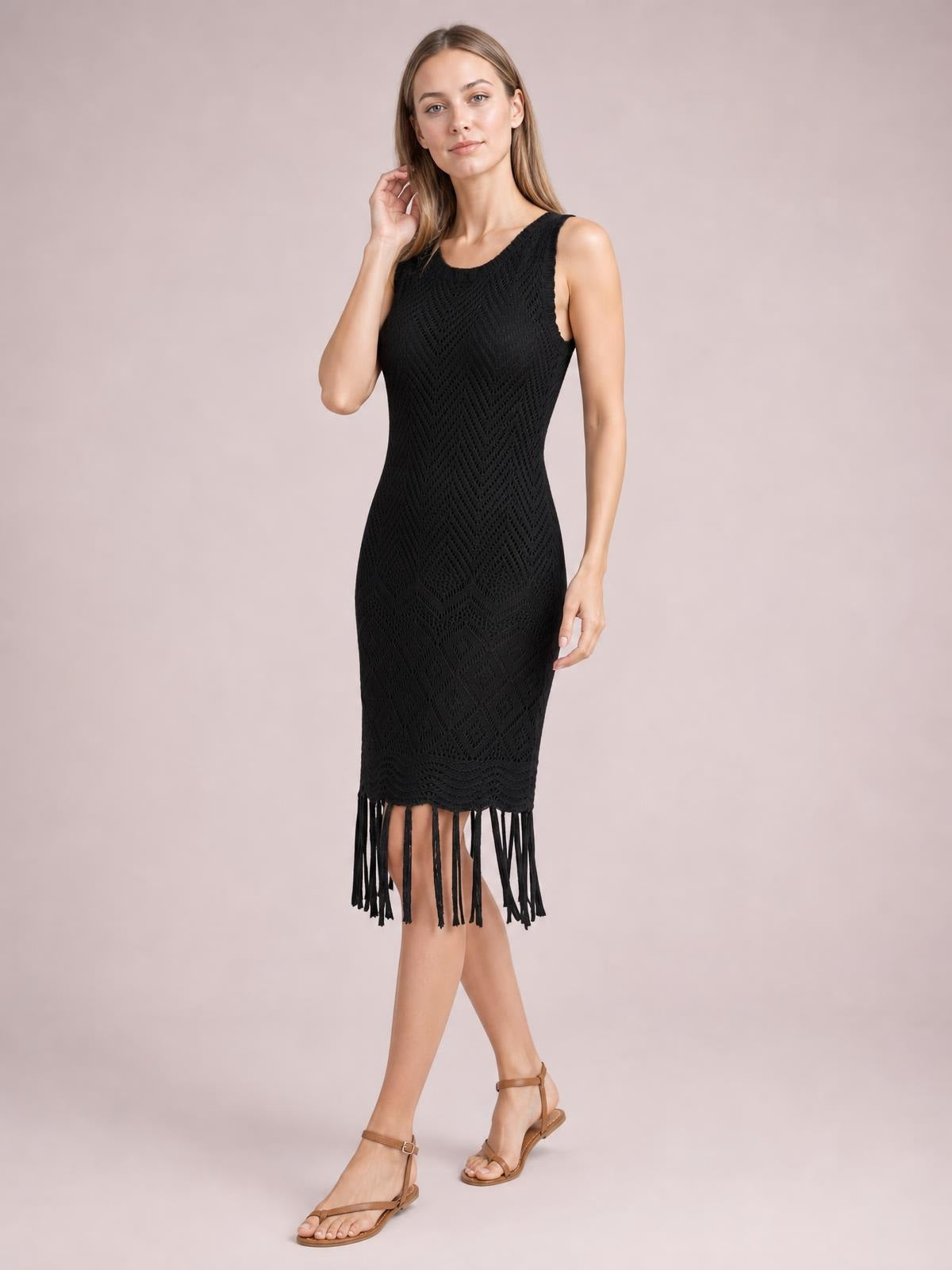 A model poses in a black sleeveless openwork cover-up dress with a fitted silhouette, textured knit construction, and long fringe trim along the hem.
