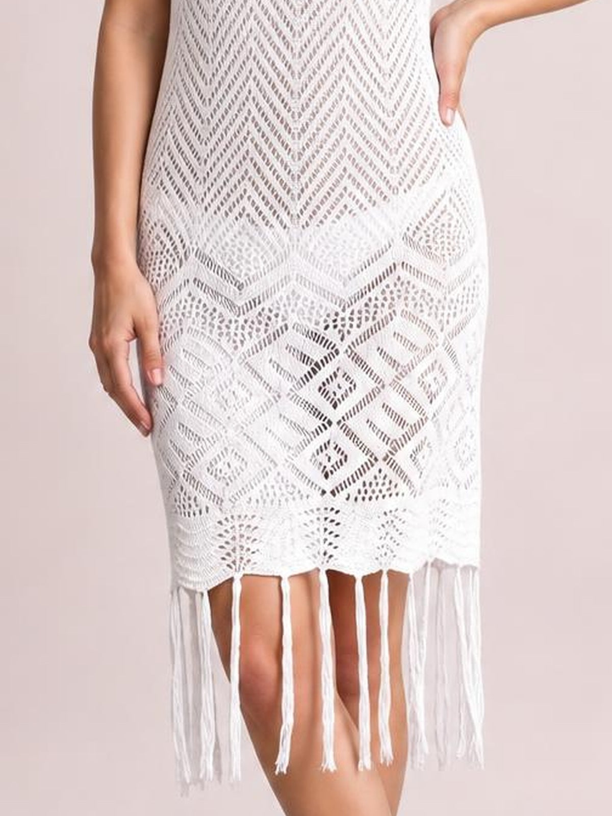 A standing model wears a white sleeveless openwork cover-up dress with a geometric chevron pattern, straight silhouette, and long fringe trim at the hem.