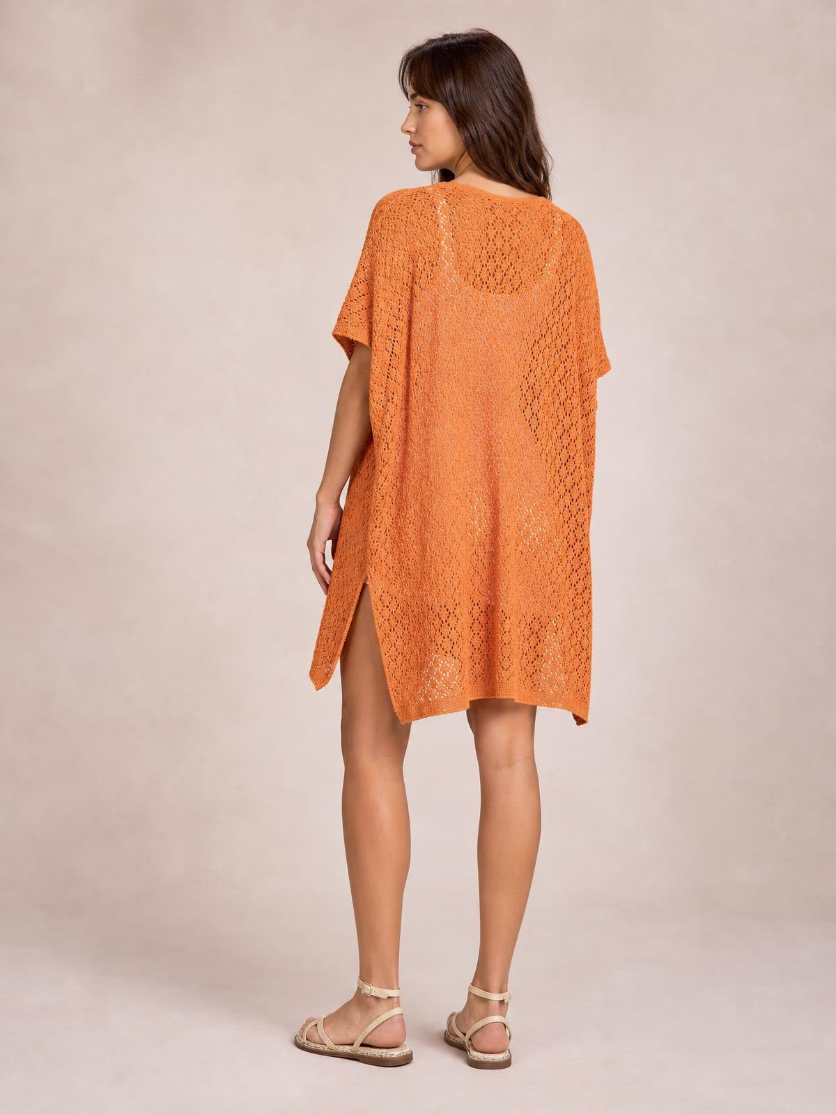 A model poses in a three-quarter back view wearing an orange semi-sheer openwork cover up with a loose silhouette, short sleeves, scoop neck, and side slit.