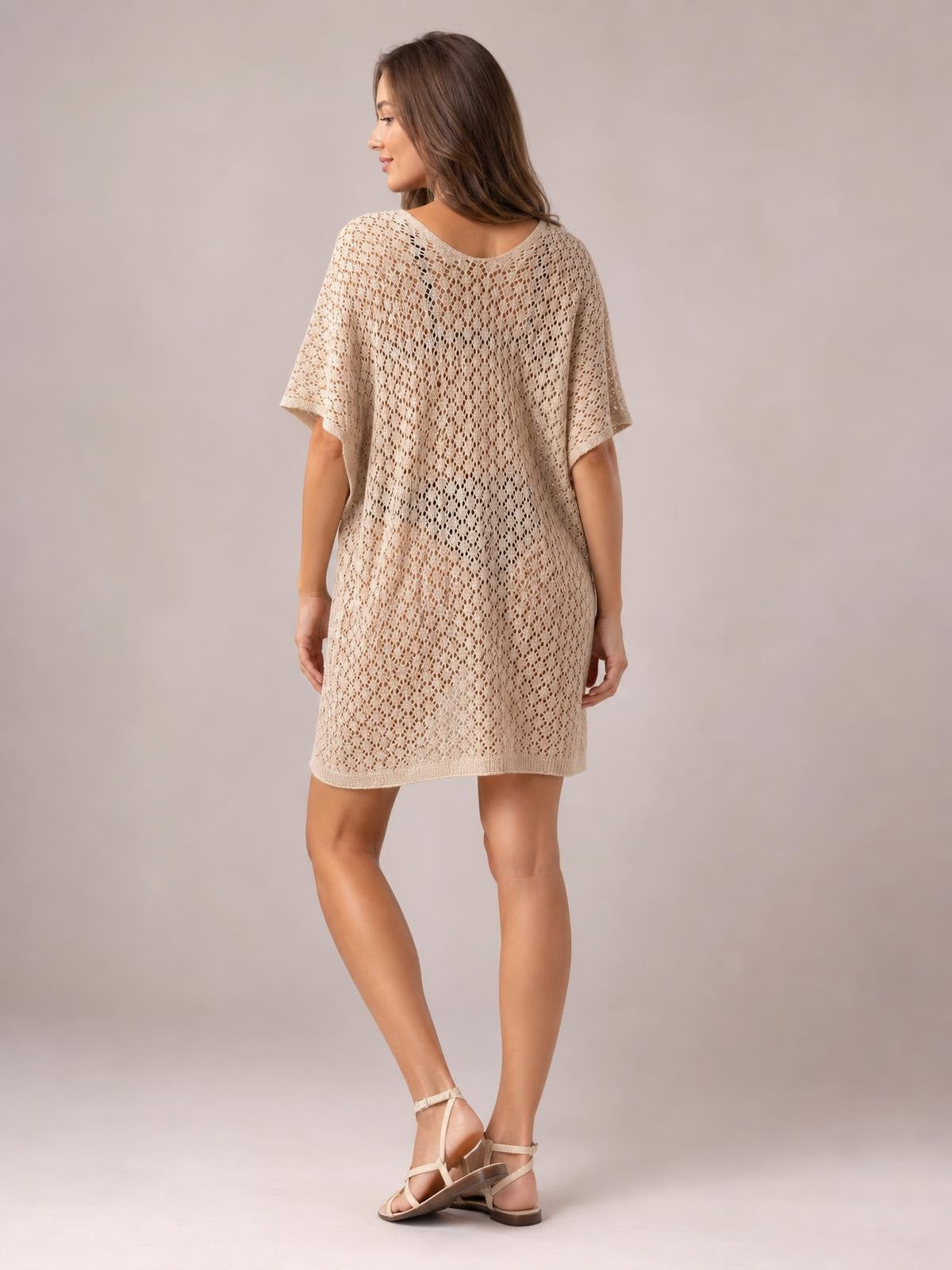 Model shown from behind in a beige semi-sheer openwork cover-up with a scoop neck, short sleeves, and a relaxed straight silhouette, standing with crossed legs.