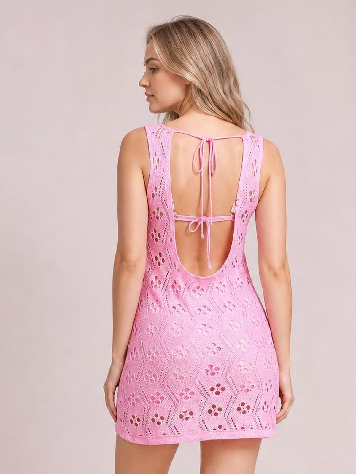 A model poses from the back in a pink semi-sheer openwork cover-up with a relaxed mini silhouette, wide straps, and tie closures framing the open back.