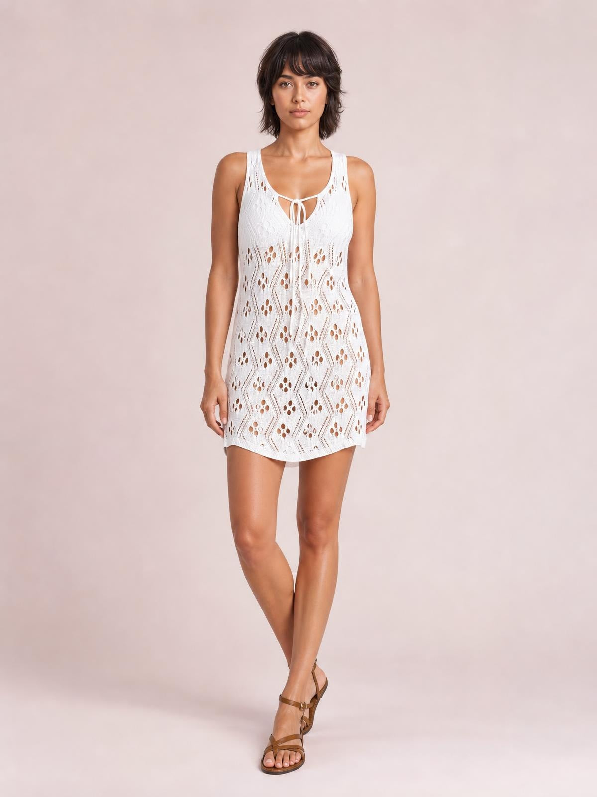 A model stands with crossed legs wearing a white semi-sheer openwork cover-up with a tie neck, scoop neckline, sleeveless cut, and relaxed mini-length silhouette.