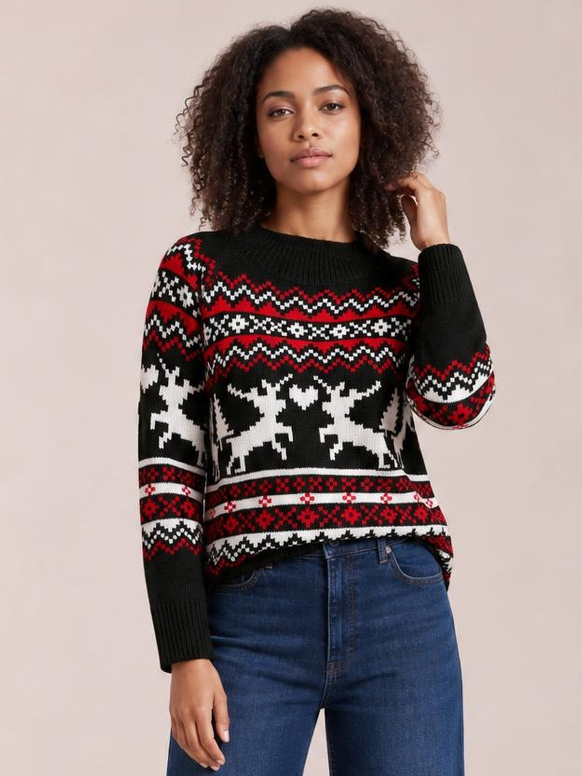 A model wears a black, red, and white reindeer round-neck long-sleeve sweater with ribbed trim and patterned knit detail, posing with one hand in her hair.