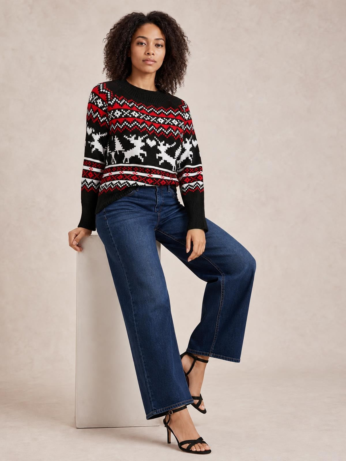 Model seated in a black reindeer sweater with a round neck, long sleeves, red and white patterned bands, and a relaxed silhouette.