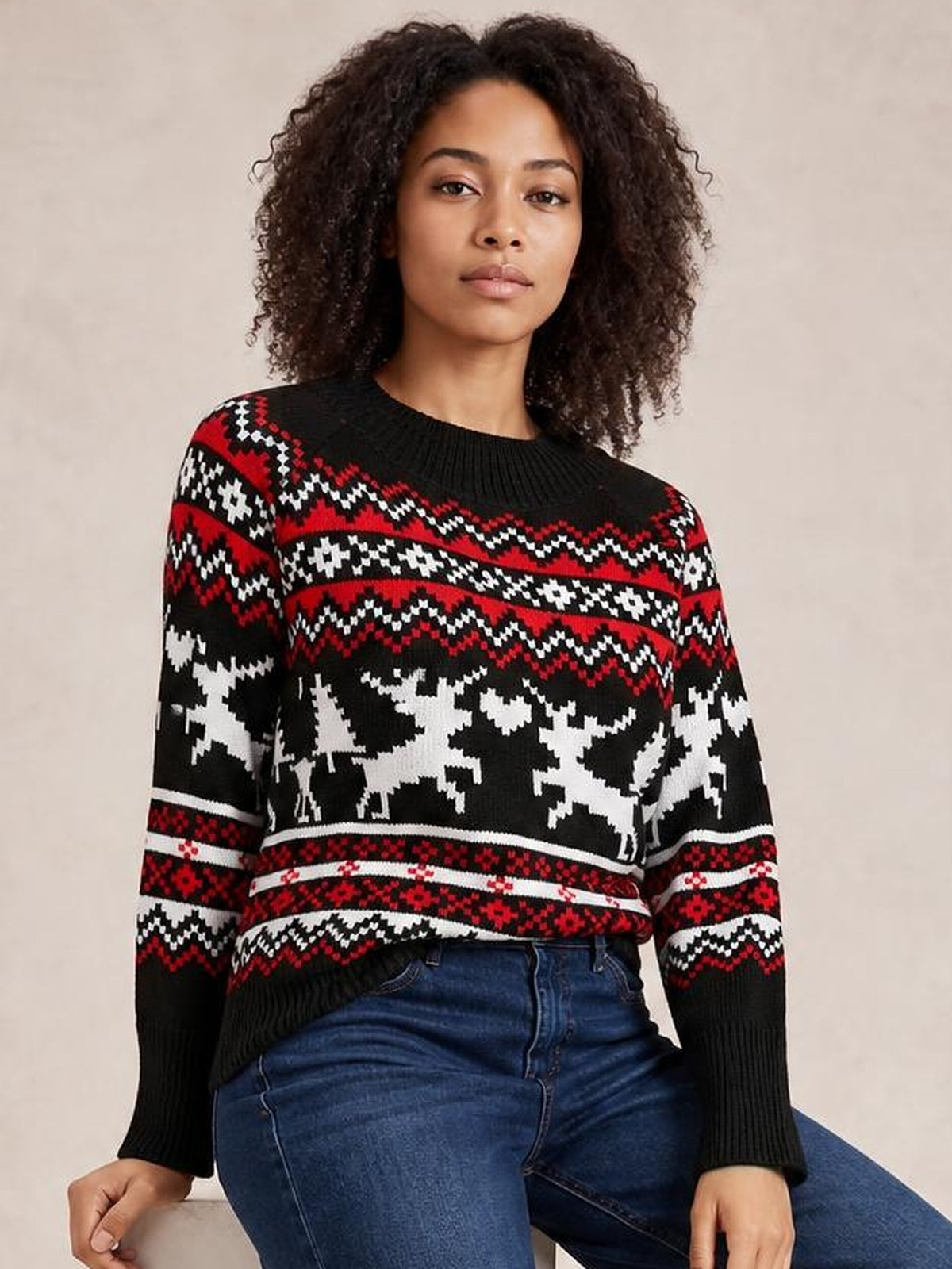 A seated model wears a black reindeer round neck long sleeve sweater with red and white patterned bands, ribbed cuffs, and a relaxed silhouette.