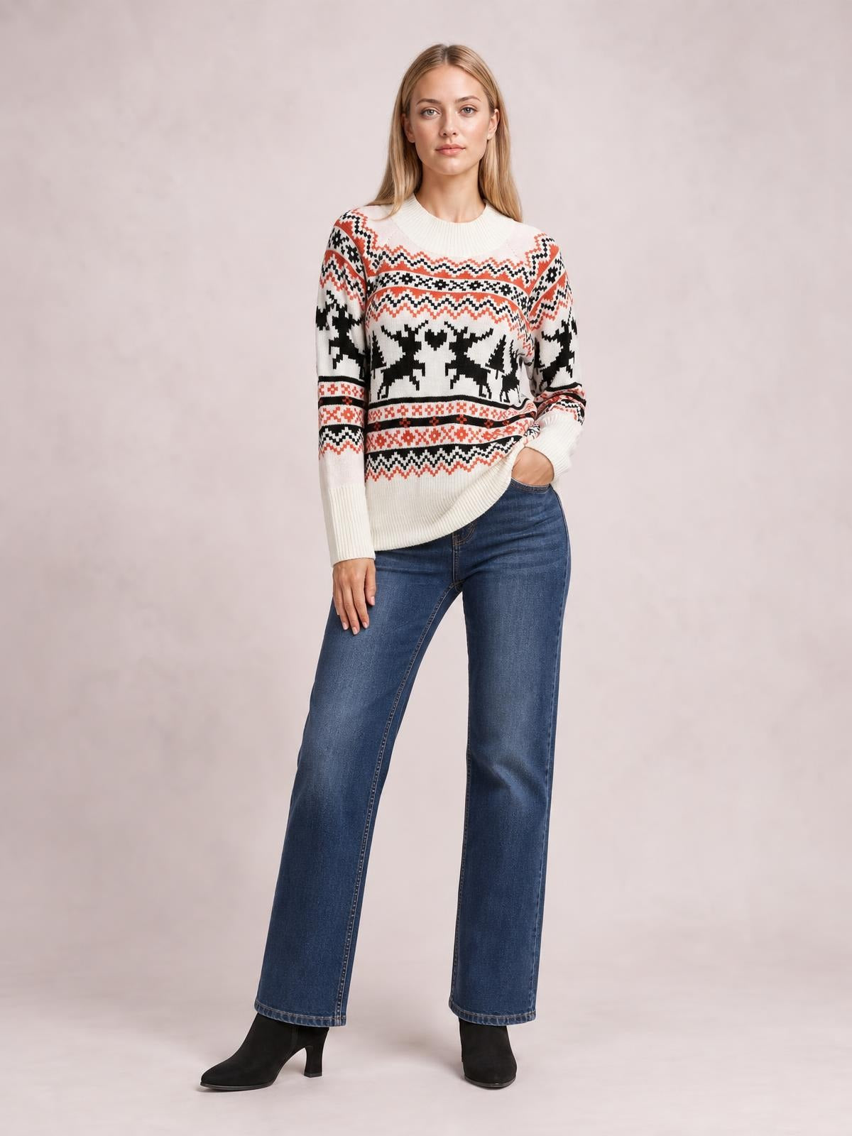 Model wearing a cream reindeer round neck long sleeve sweater with black and red patterned bands, ribbed cuffs and hem, posed with one hand in her jeans pocket.