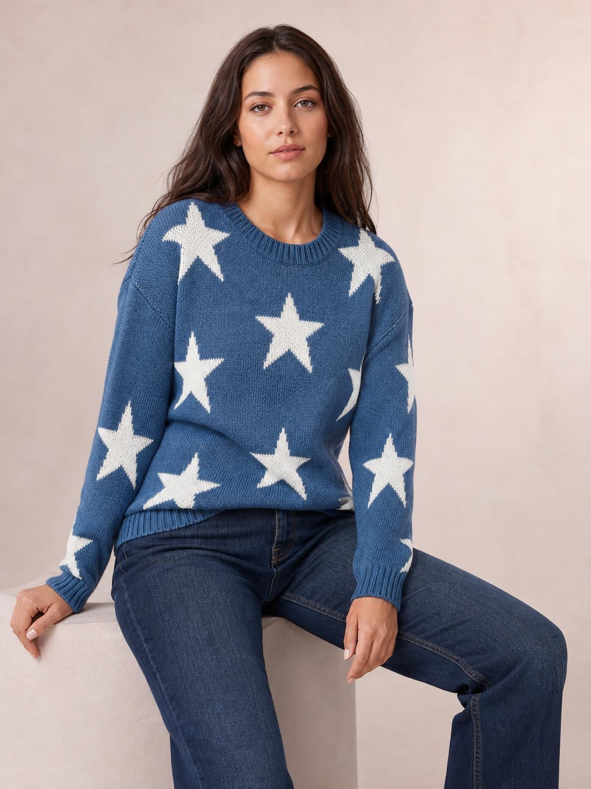 Model seated in a blue round neck dropped shoulder sweater with white star motifs, ribbed neckline, cuffs and hem, and a relaxed silhouette.
