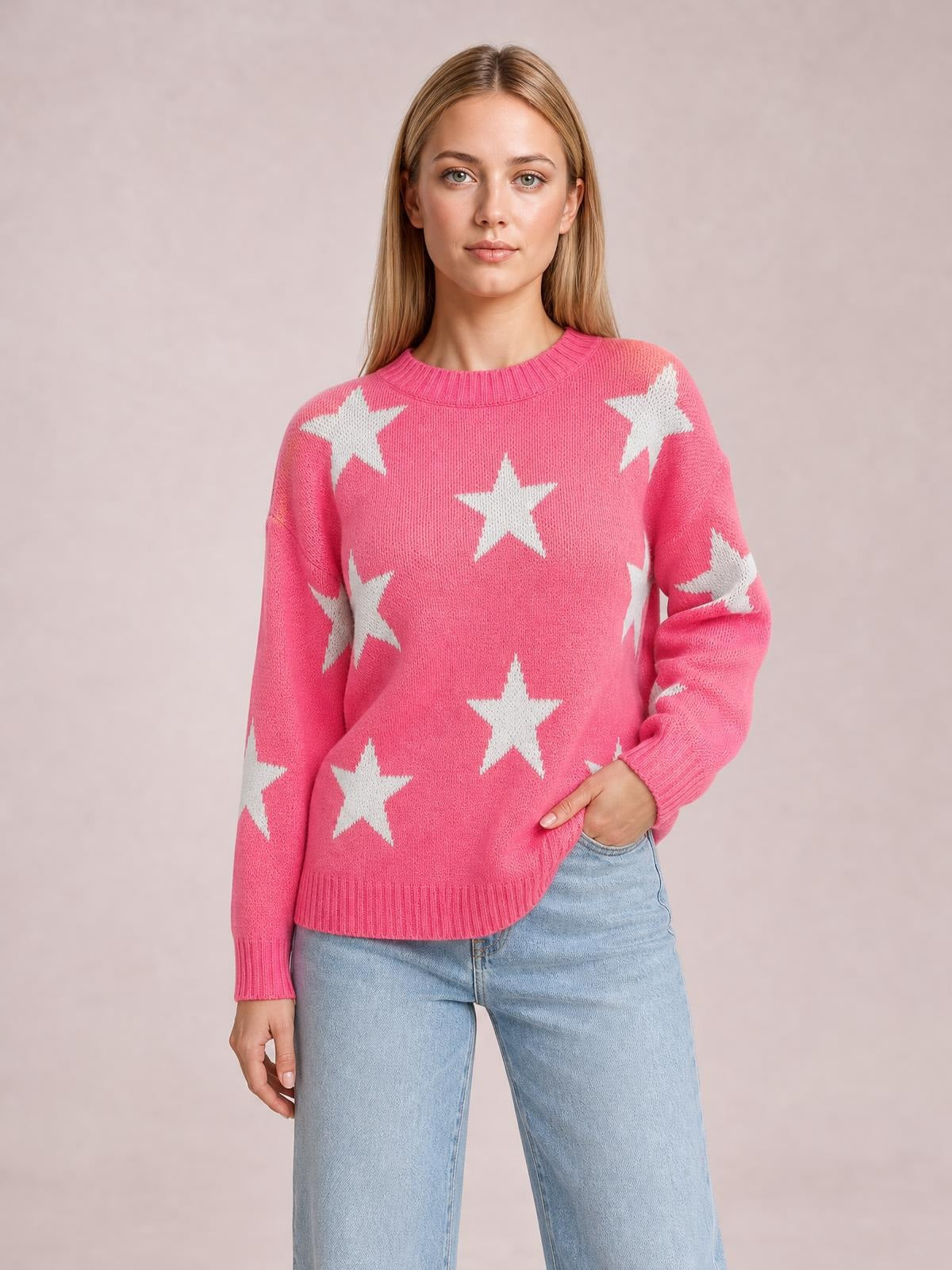 Model poses in a pink round neck dropped shoulder sweater with white star motifs, ribbed cuffs and hem, and a relaxed silhouette, one hand in her jeans pocket.
