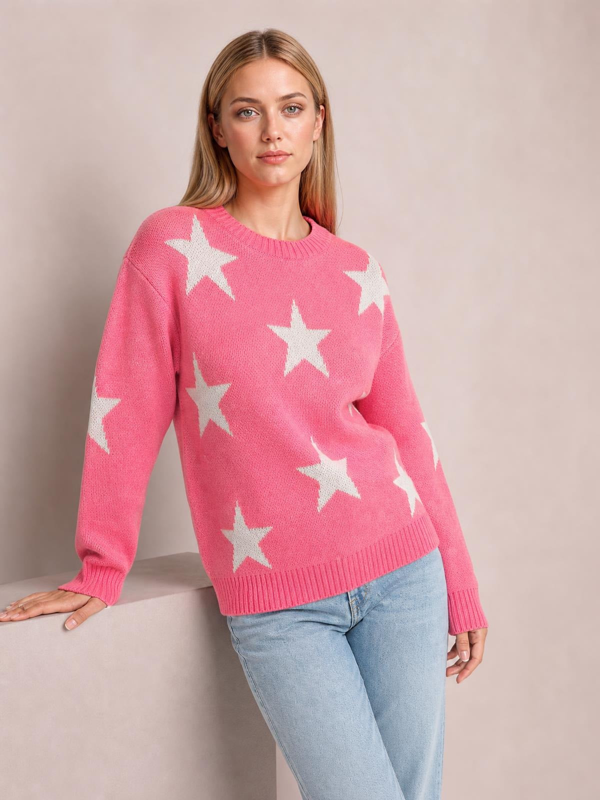 Model leans with one hand supported, wearing a pink star-pattern round neck sweater with dropped shoulders, long sleeves, ribbed cuffs and hem, and a relaxed silhouette.