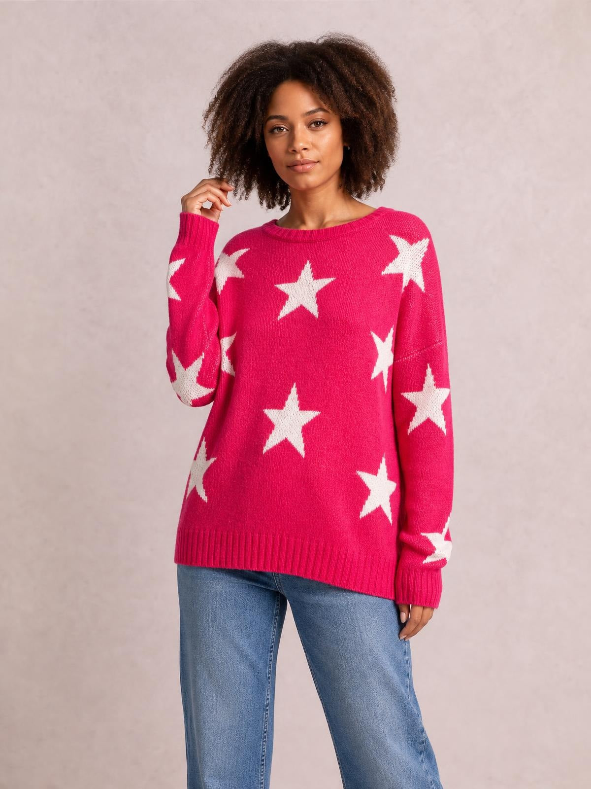 Model poses with one hand near her hair in a bright pink dropped-shoulder sweater featuring a round neck, white star pattern, long sleeves, and ribbed trims.